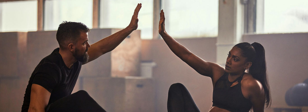 Man and woman in workout gear high-fiving each other on the floor during a gym session.