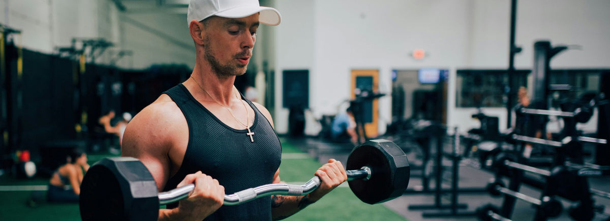 Athletic man wearing a black tank top and cap doing EZ-bar bicep curls in a gym.