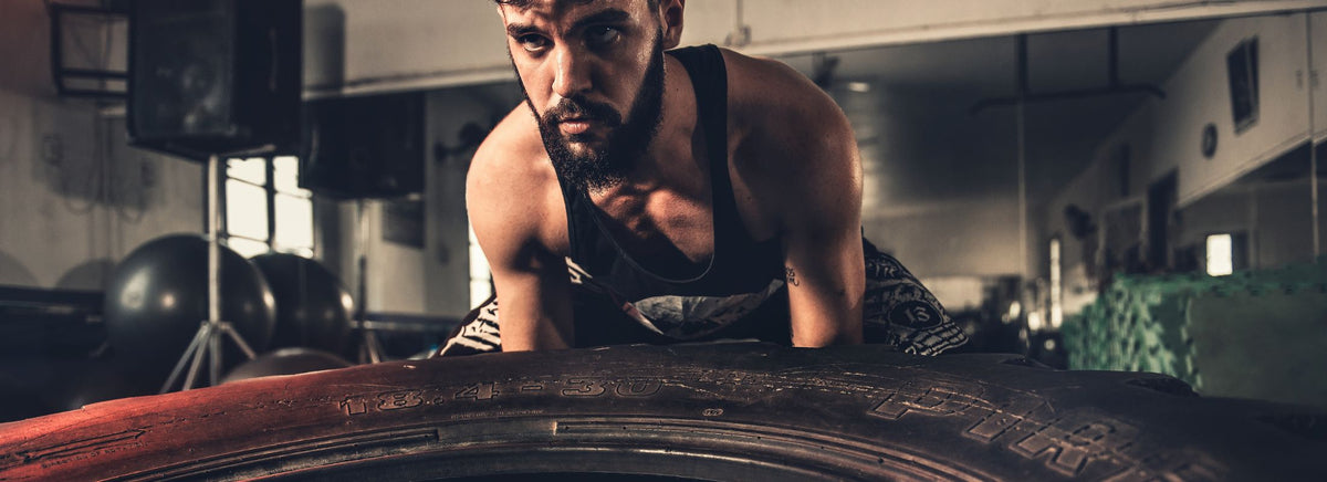 Focused athletic man in a tank top leaning over a large gym tire.