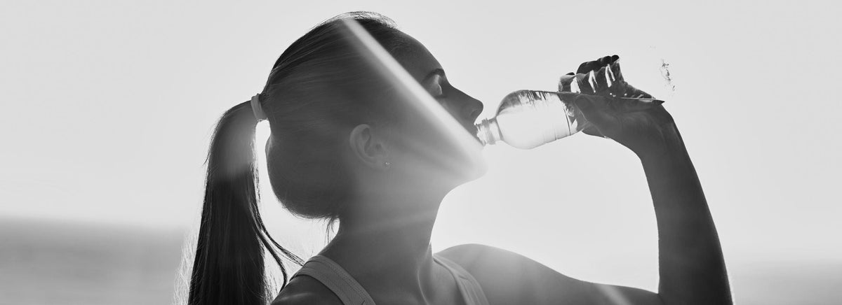 Silhouette of a woman drinking water from a plastic bottle outdoors with sunlight glowing behind her.