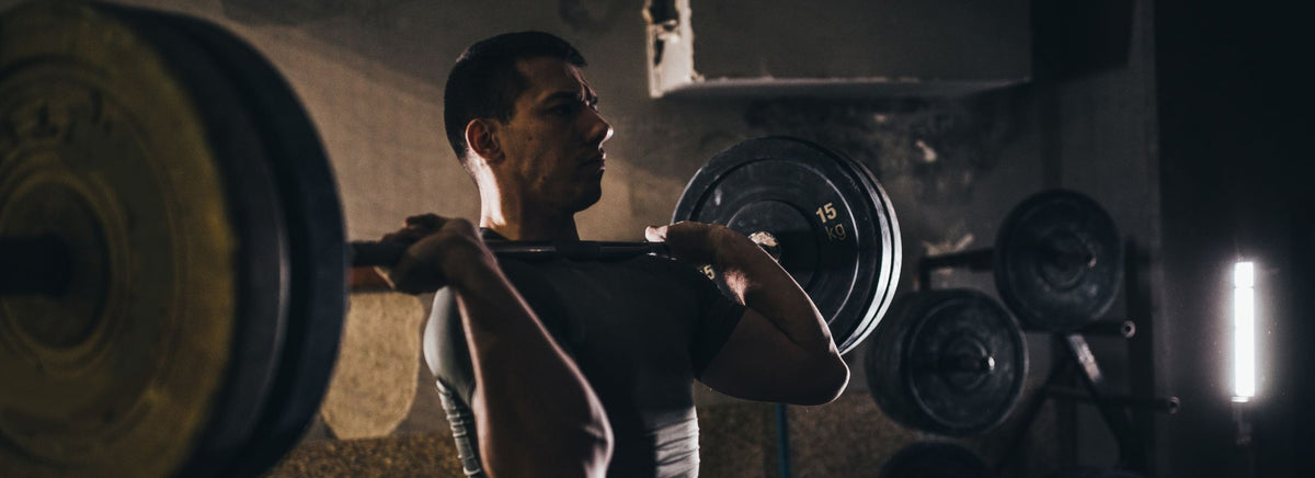 Shirtless muscular man performing an overhead barbell lift in a gym.