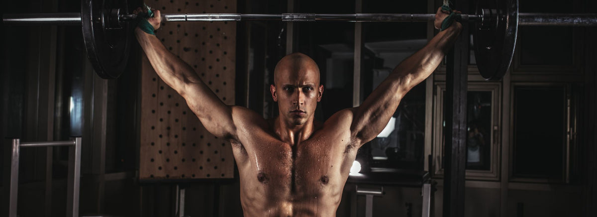 Shirtless muscular man performing an overhead barbell lift in a gym.