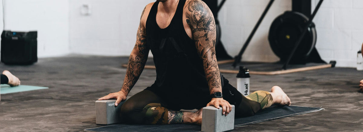 Tattooed person in black gym wear performing a deep split leg stretch using yoga blocks on a mat.