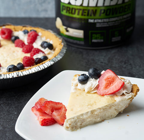 Slice of cheesecake topped with strawberries and blueberries next to a full pie and protein powder tub.
